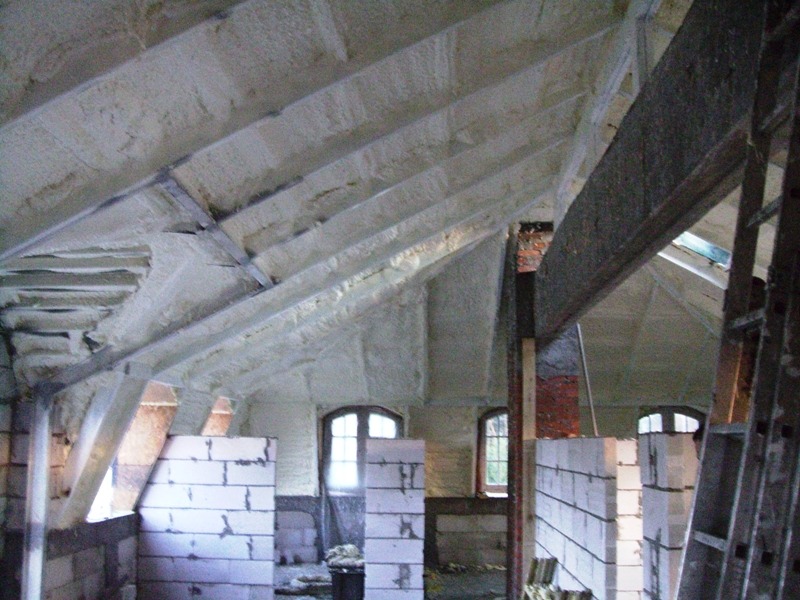 Poddasze z oknami po natrysku pianki izolacyjnej, widoczne elementy konstrukcyjne i fragmenty muru z białych bloczków. Izolacja między krokwiami dachu.