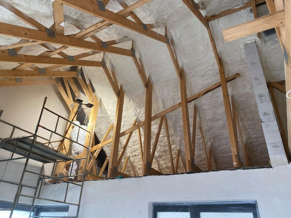 Skośne poddasze z drewnianą konstrukcją pokryte białą pianką izolacyjną, widok z dołu. Pracownik w kombinezonie na rusztowaniu.