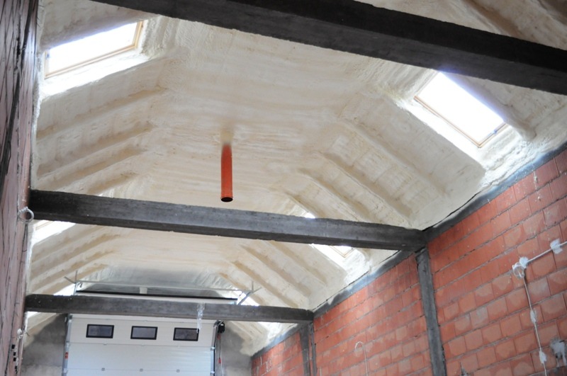 Poddasze z oknami dachowymi po natrysku pianką, widoczne drewniane belki konstrukcyjne i fragment muru z czerwonej cegły. W tle brama garażowa.