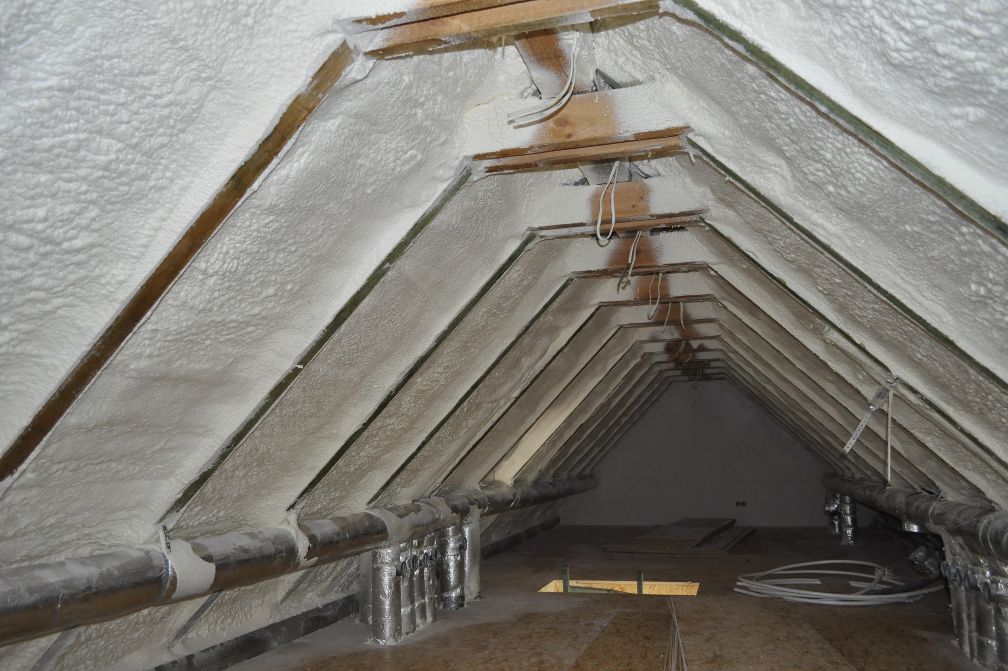 Poddasze po ociepleniu pianką PUR, widoczne krokwie i instalacja wentylacyjna. Biała piana szczelnie wypełnia przestrzeń, tworząc jednolitą warstwę izolacyjną.