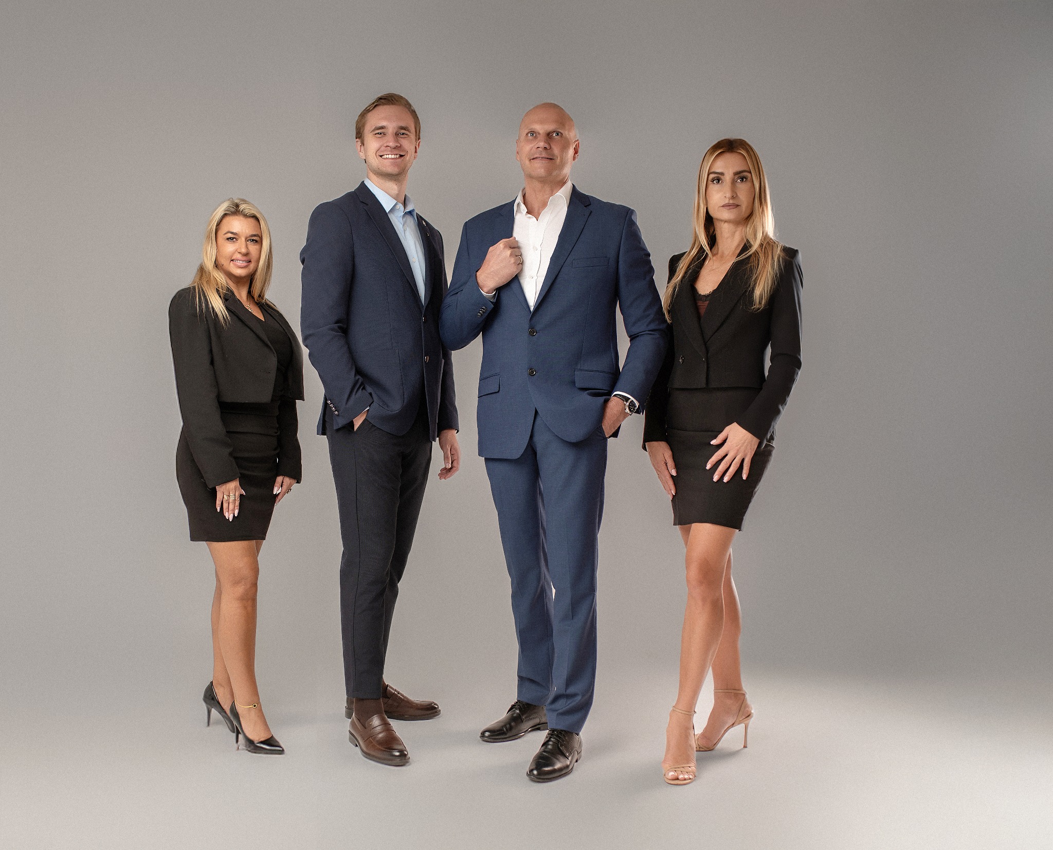 Zespół agentów nieruchomości w garniturach pozuje na szarym tle. Mężczyzna w środku rozluźnia kołnierz. Profesjonalny wizerunek firmy.