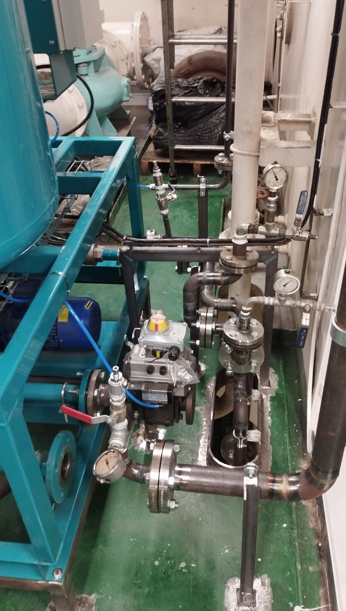 Kompleksowa instalacja przemysłowa z rurami, zaworami i manometrami w hali. Widoczny silnik elektryczny i zbiornik na ramie. Elementy spawane.