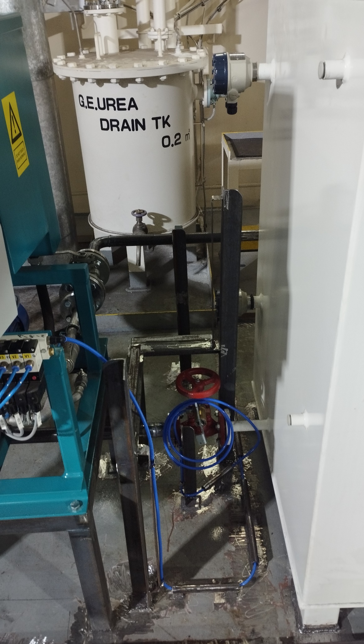 Fragment instalacji przemysłowej z zbiornikiem 'G.E.UREA DRAIN TK 0.2 m³'. Widoczne elementy konstrukcji stalowej, rury i zawory. Spawy na podłodze.