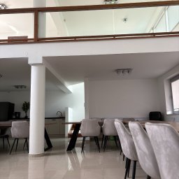 Twoja Sala Eventowa - Przestronne wnętrze sali konferencyjnej z długim stołem, krzesłami z szarego weluru i widokiem na antresolę z drewnianą balustradą. Jasne płytki podłogowe.