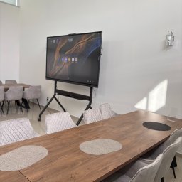 Twoja Sala Eventowa - Sala konferencyjna z dużym ekranem dotykowym na mobilnym stojaku, stół z szarymi podkładkami i krzesłami. Oświetlenie dzienne.