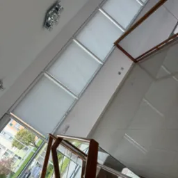 Nowoczesne rolety okienne w jasnym wnętrzu z drewnianymi elementami balustrady i widokiem na zewnątrz. Oświetlenie sufitowe.