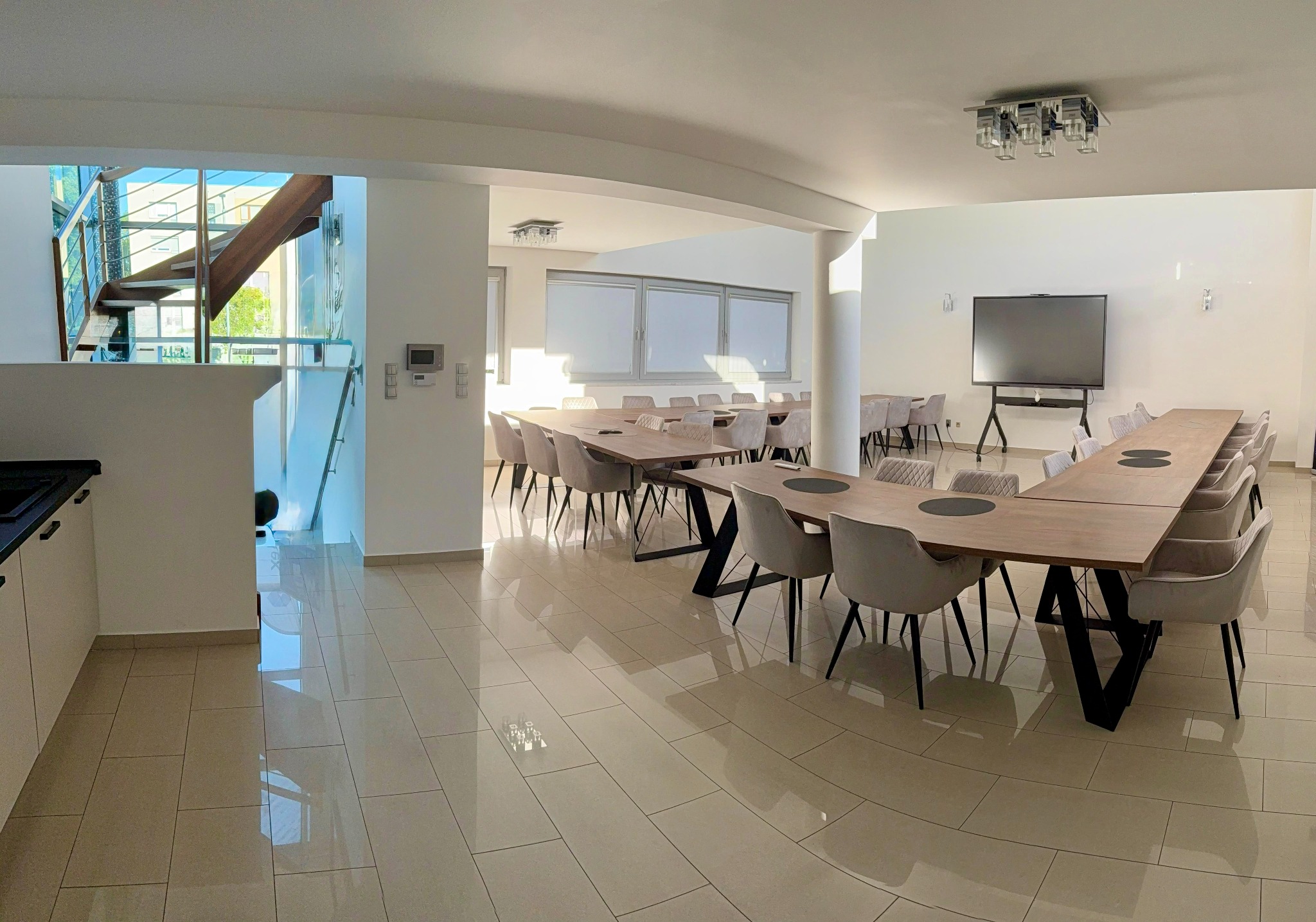 Nowoczesna sala konferencyjna z długimi stołami, krzesłami i dużym ekranem, gotowa na event. Jasne wnętrze z widocznymi schodami i elementami wystroju.