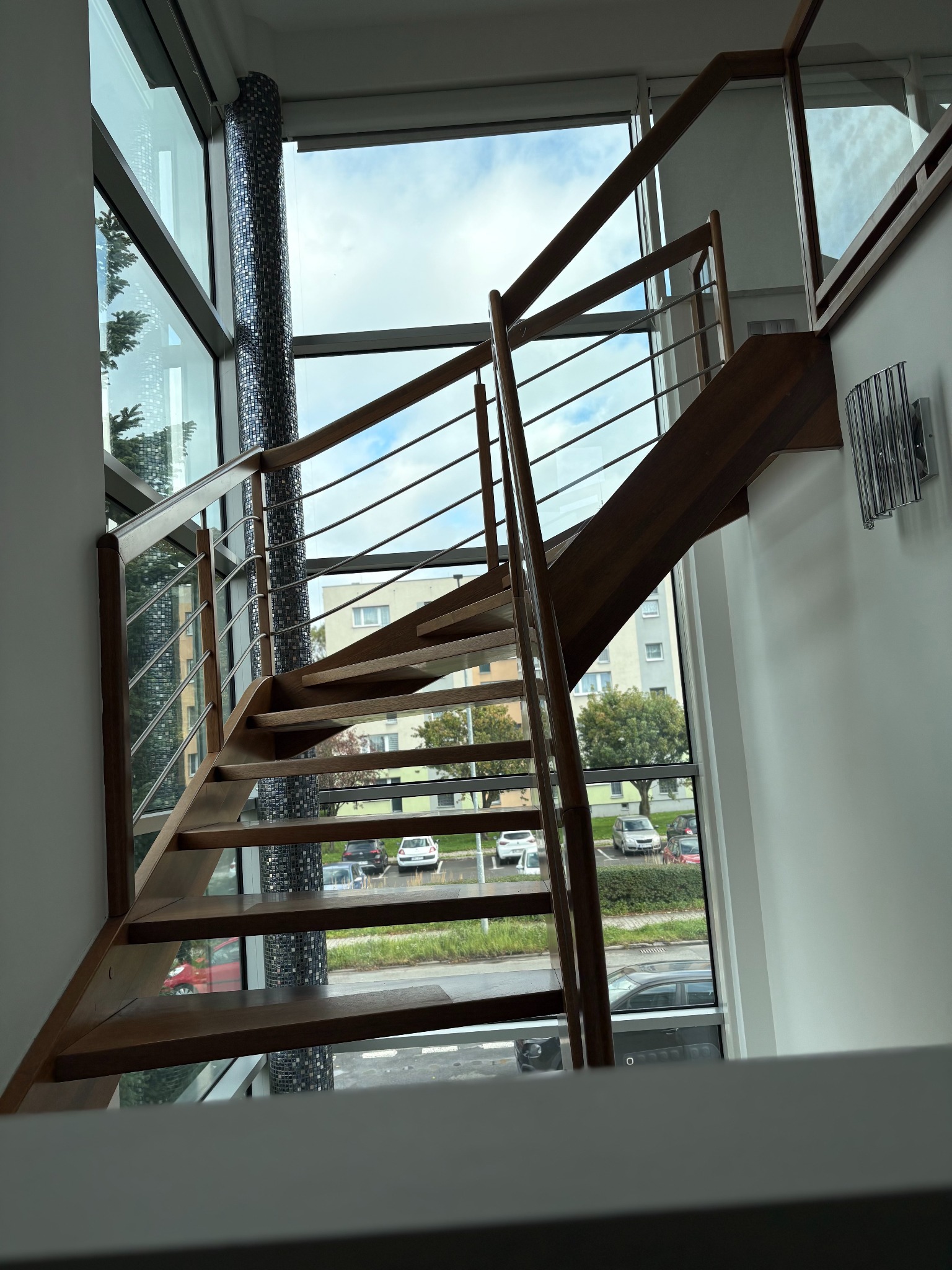 Drewniane schody z metalowymi barierkami w nowoczesnym wnętrzu z wysokimi oknami i mozaikową kolumną. Widok na zewnątrz z samochodami i budynkami.