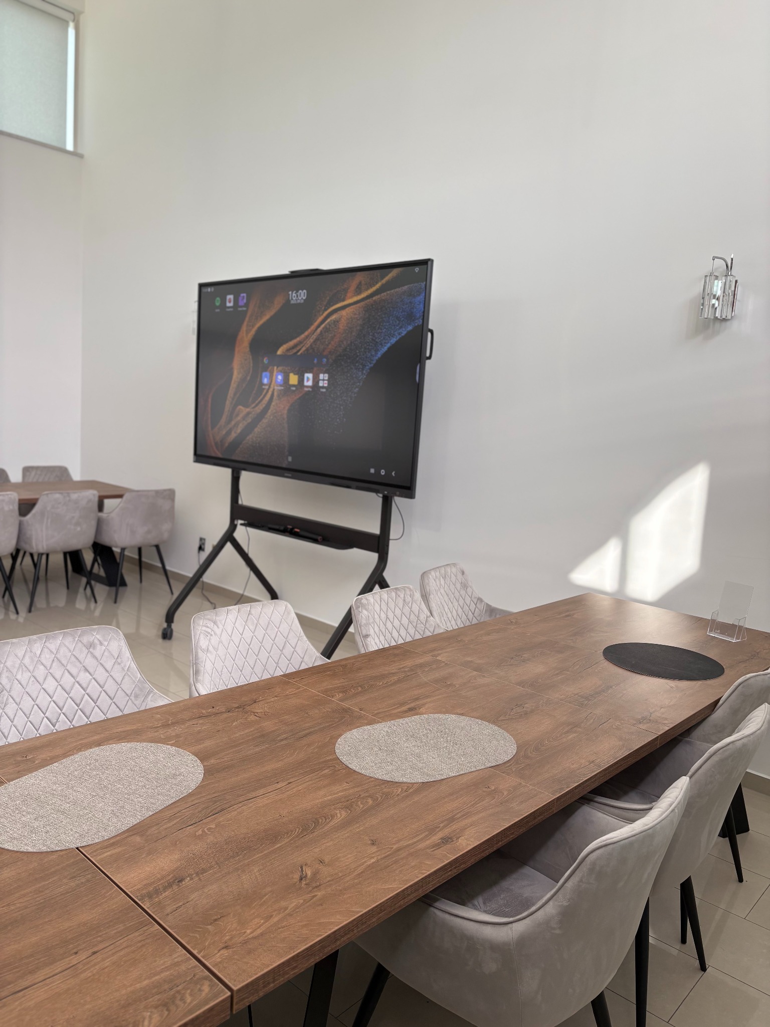 Sala konferencyjna z dużym ekranem dotykowym na mobilnym stojaku, stół z szarymi podkładkami i krzesłami. Oświetlenie dzienne.
