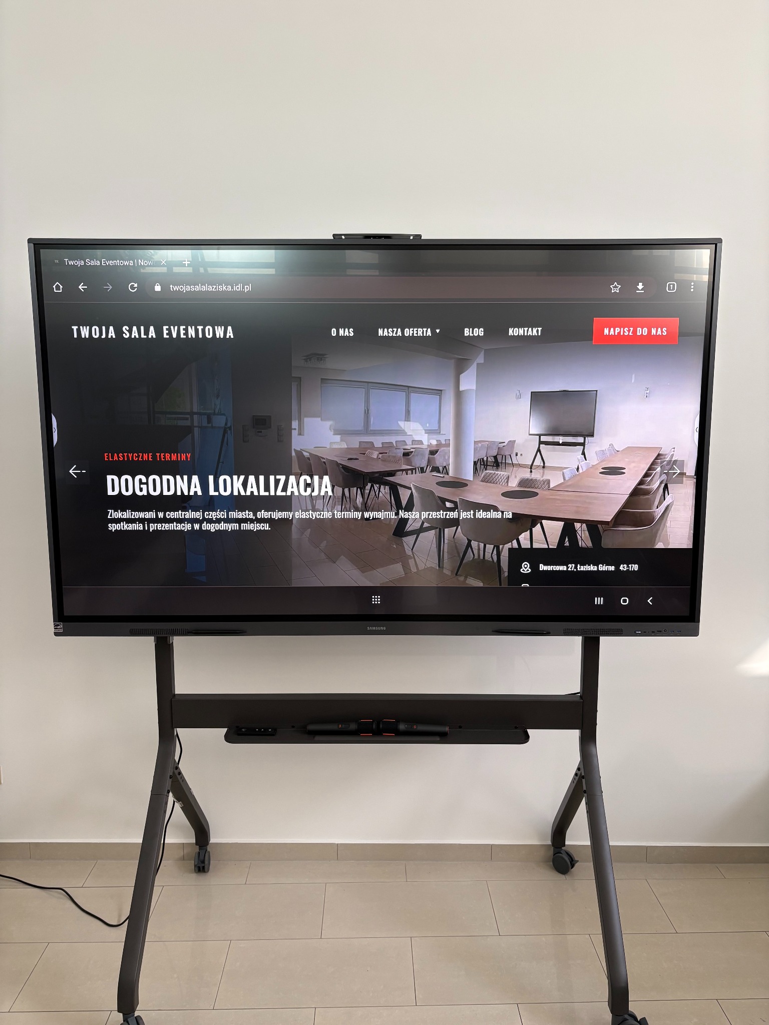 Interaktywny monitor Samsung na stojaku prezentuje stronę internetową sali eventowej z ofertą wynajmu przestrzeni na spotkania i prezentacje. Widoczny adres i dogodna lokalizacja.