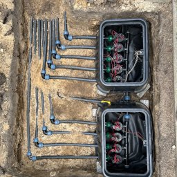 Krzysztof Garstka - Fragment systemu nawadniania w wykopie ziemnym, z widocznymi rurami, złączkami i dwoma czarnymi skrzynkami sterującymi, częściowo zasypanymi ziemią.