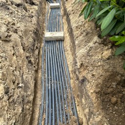 Krzysztof Garstka - Wkopane w ziemię rury systemu nawadniania, zabezpieczone betonowymi płytkami, z widocznymi czarnymi skrzynkami na początku instalacji.