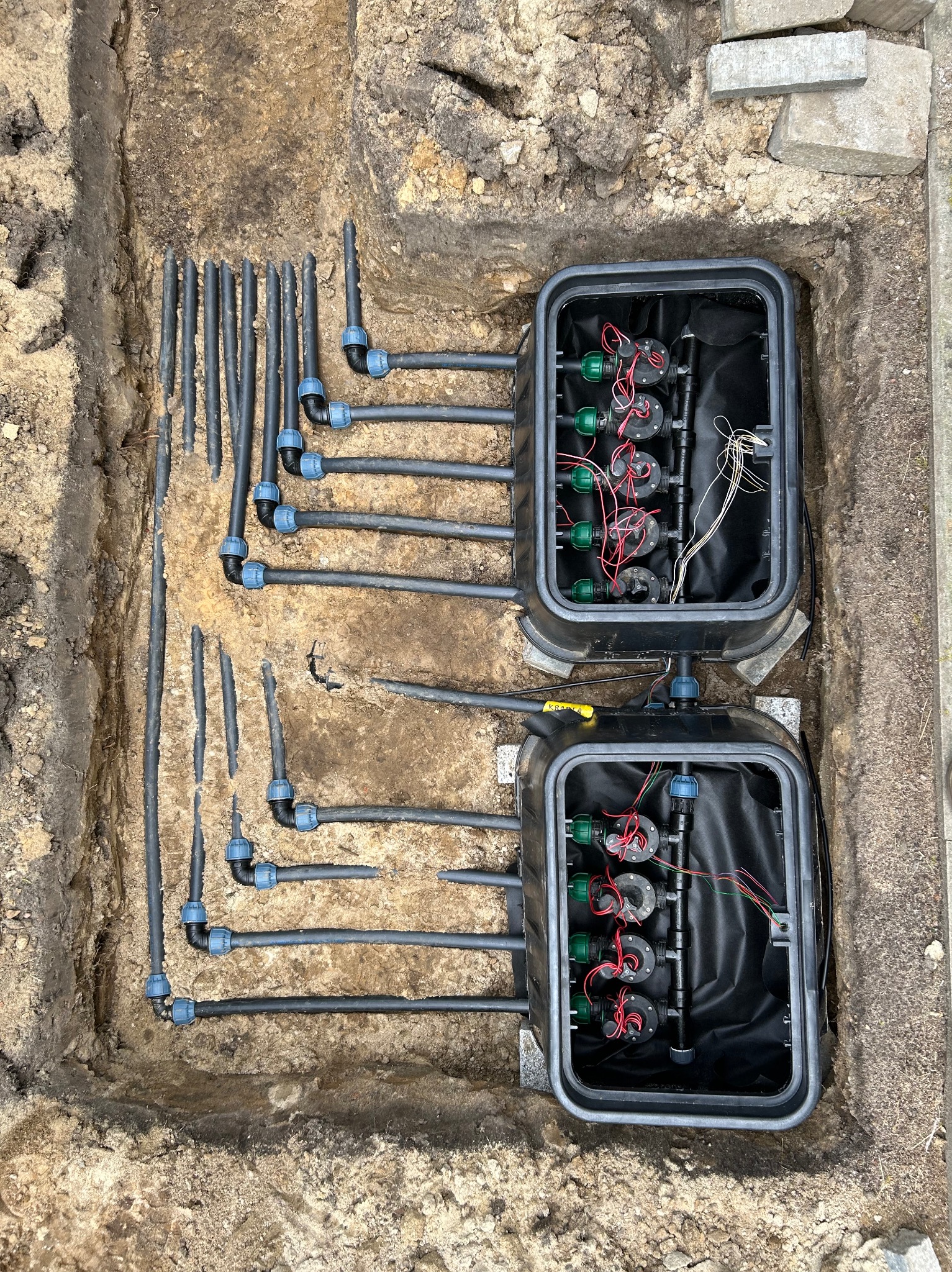 Fragment systemu nawadniania w wykopie ziemnym, z widocznymi rurami, złączkami i dwoma czarnymi skrzynkami sterującymi, częściowo zasypanymi ziemią.