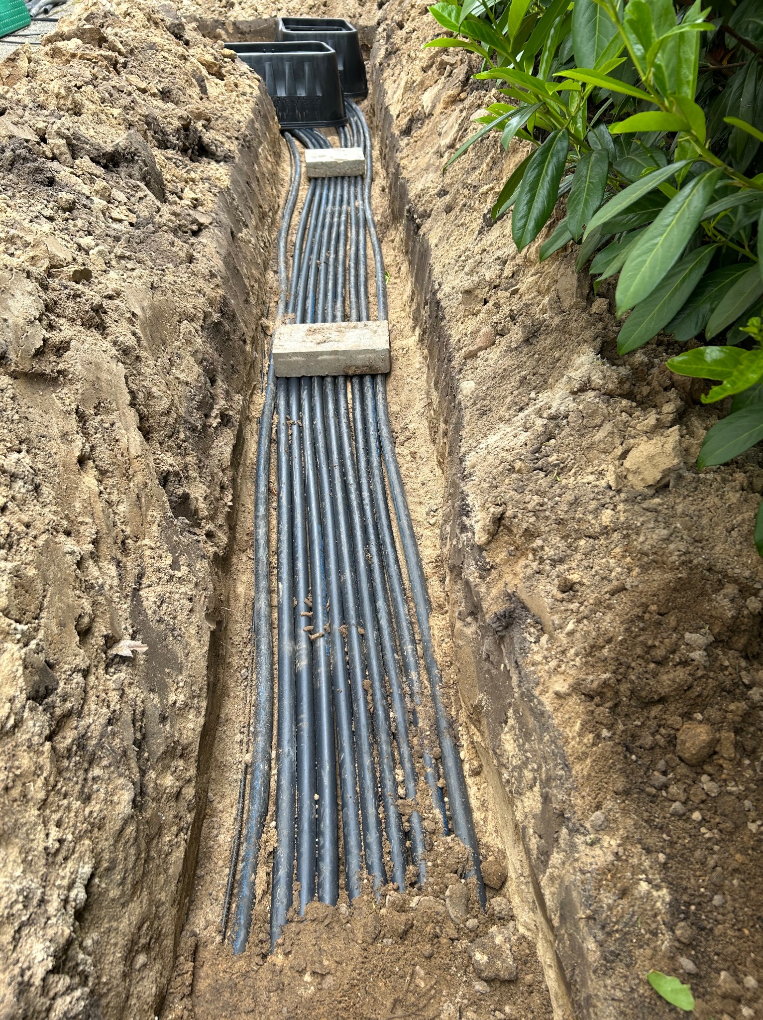 Wkopane w ziemię rury systemu nawadniania, zabezpieczone betonowymi płytkami, z widocznymi czarnymi skrzynkami na początku instalacji.