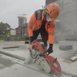 Marcin Gut Merkosmik - Pracownik w pomarańczowej kurtce przecina betonową powierzchnię szlifierką kątową Stihl, z widocznymi budynkami w tle i pyłem powstającym podczas cięcia.
