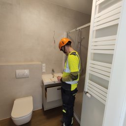 Marcin Gut Merkosmik - Instalator w kasku montuje umywalkę w nowoczesnej łazience z kabiną prysznicową i grzejnikiem drabinkowym. Widoczne płytki w odcieniach beżu i drewnopodobna podłoga.