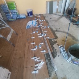 Marcin Gut Merkosmik - Układanie drewnopodobnych płytek podłogowych z użyciem klipsów poziomujących, widoczne narzędzia i materiały budowlane w pomieszczeniu w trakcie prac glazurniczych.