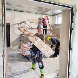 Marcin Gut Merkosmik - Ekipa w trakcie prac szpachlarskich: mężczyzna z uśmiechem trzyma płytę z nałożoną szpachlą, w tle rusztowanie i współpracownicy. Widoczne worki ze szpachlą.