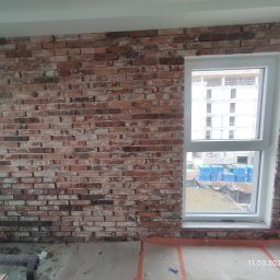 Marcin Gut Merkosmik - Ściana z cegły klinkierowej wewnątrz pomieszczenia, z oknem. Widoczne narzędzia i materiały budowlane, sugerujące etap wykończeniowy. Nietypowe ułożenie cegieł.