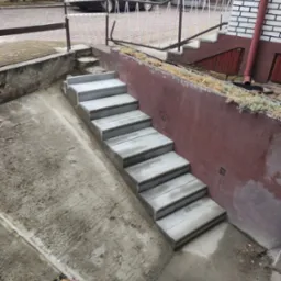 Nowe, szare schody betonowe prowadzą w dół, kontrastując z czerwoną ścianą i elementami zieleni. W tle widoczny budynek i samochód.