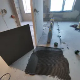 Przygotowanie do ułożenia ciemnej płytki podłogowej z nałożonym klejem w paski. W tle widoczne narzędzia i materiały budowlane oraz fragment ułożonej podłogi z krzyżykami dystansowymi.