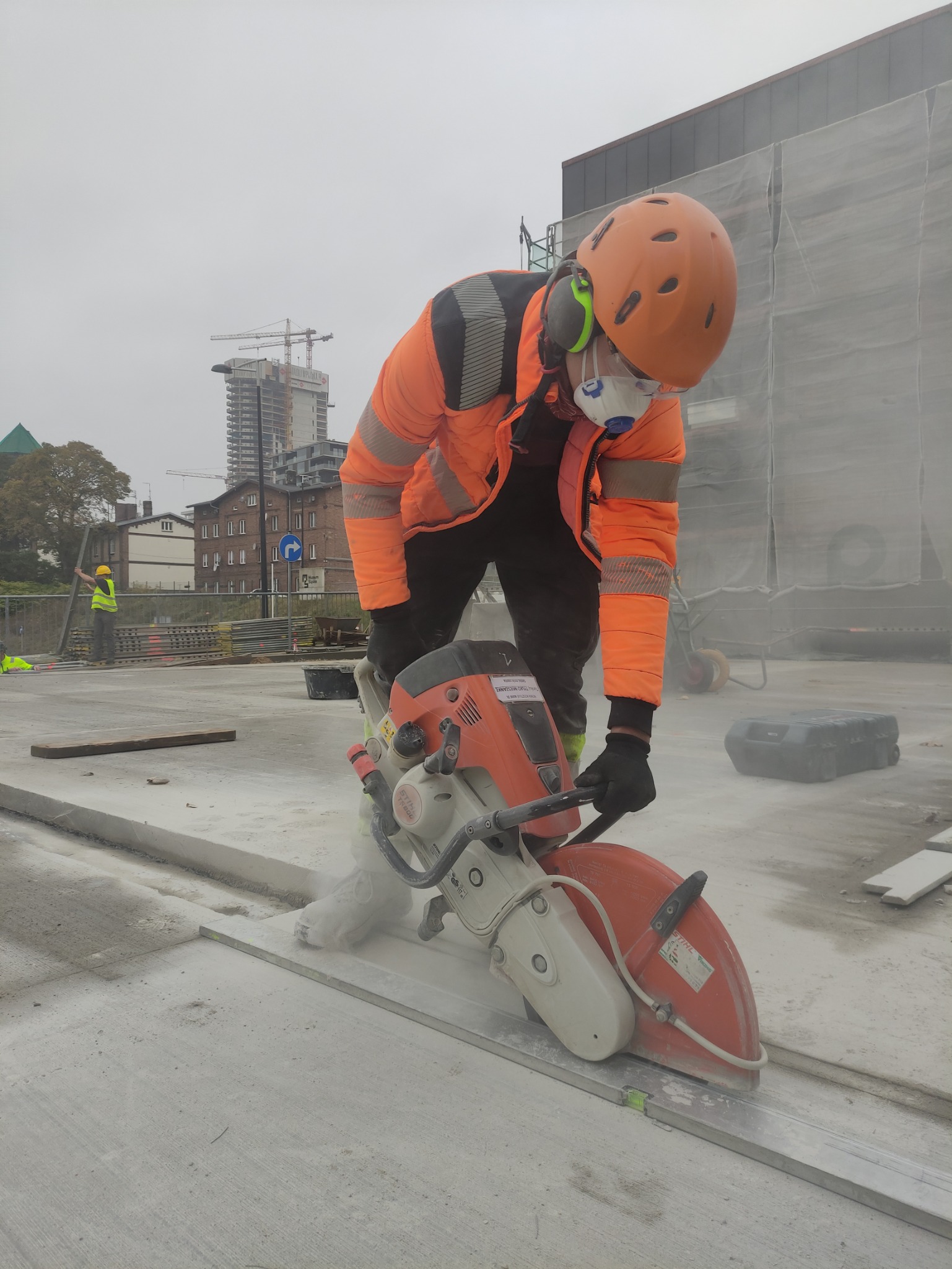 Pracownik w pomarańczowej kurtce przecina betonową powierzchnię szlifierką kątową Stihl, z widocznymi budynkami w tle i pyłem powstającym podczas cięcia.