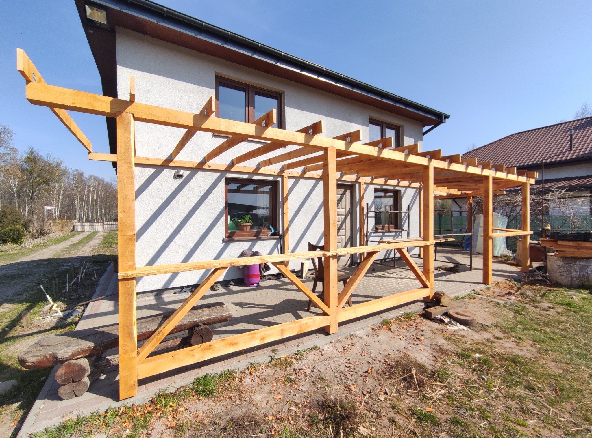 Drewniana pergola przy domu jednorodzinnym w słoneczny dzień. Konstrukcja z jasnego drewna, widoczne połączenia śrubowe. Tło: trawnik, elewacja domu z oknami.