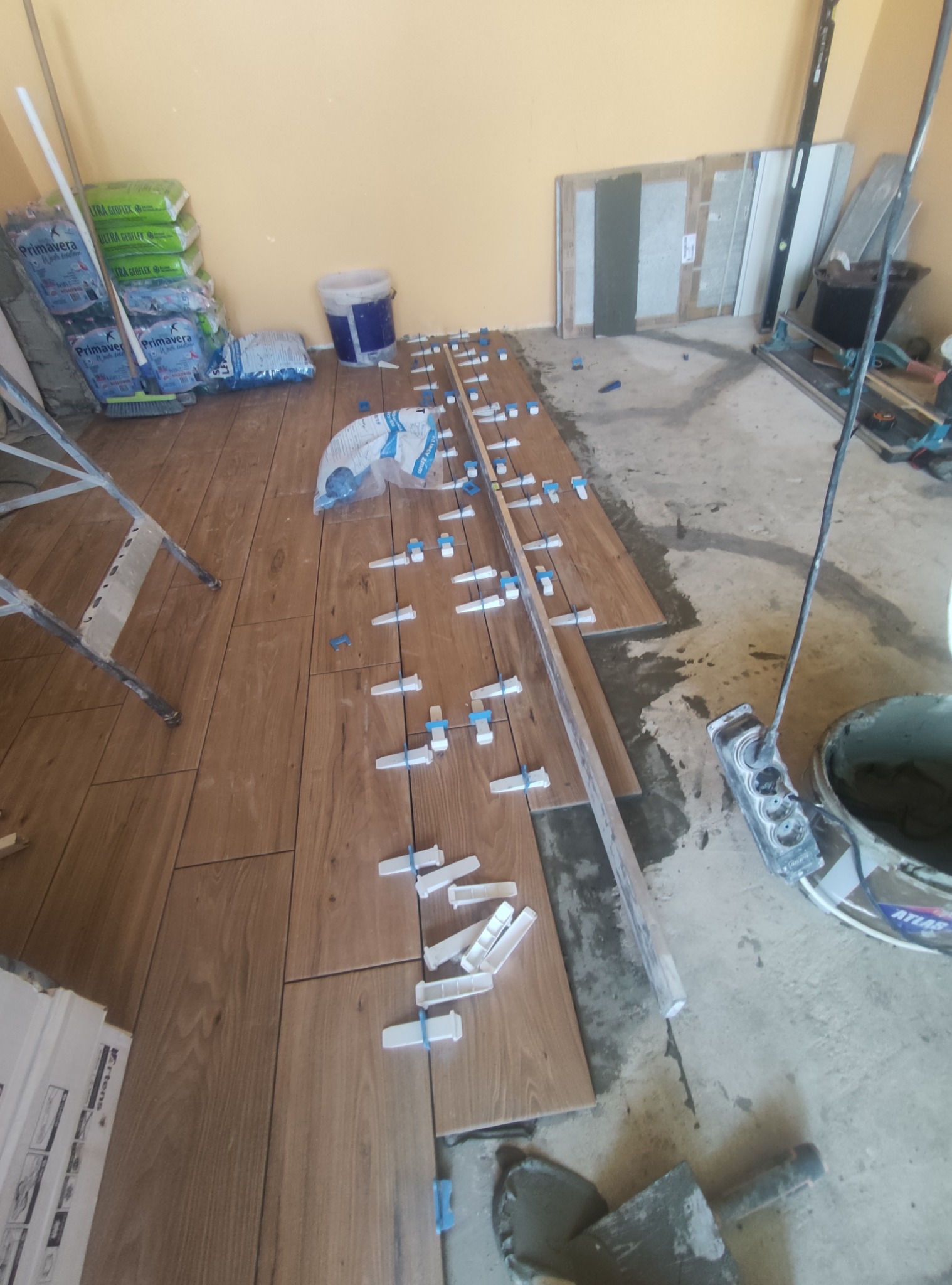 Układanie drewnopodobnych płytek podłogowych z użyciem klipsów poziomujących, widoczne narzędzia i materiały budowlane w pomieszczeniu w trakcie prac glazurniczych.