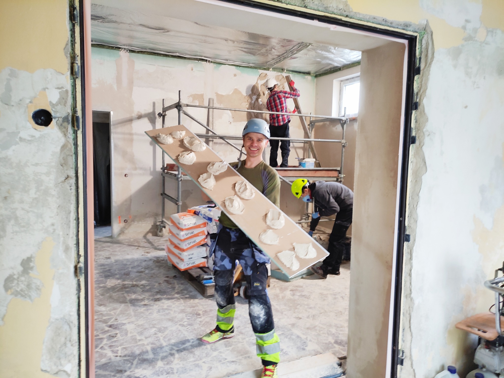 Ekipa w trakcie prac szpachlarskich: mężczyzna z uśmiechem trzyma płytę z nałożoną szpachlą, w tle rusztowanie i współpracownicy. Widoczne worki ze szpachlą.