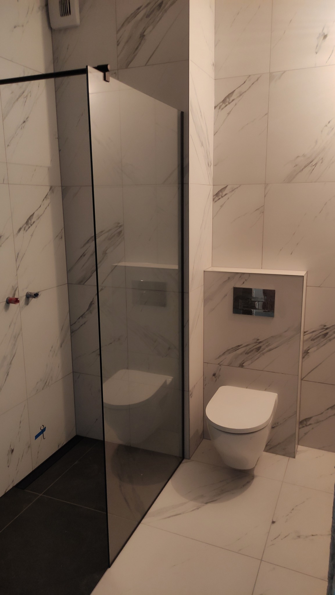 Nowoczesna łazienka z kabiną prysznicową typu walk-in i podwieszaną toaletą. Białe płytki z marmurowym wzorem. Minimalistyczny design, czarne akcenty.