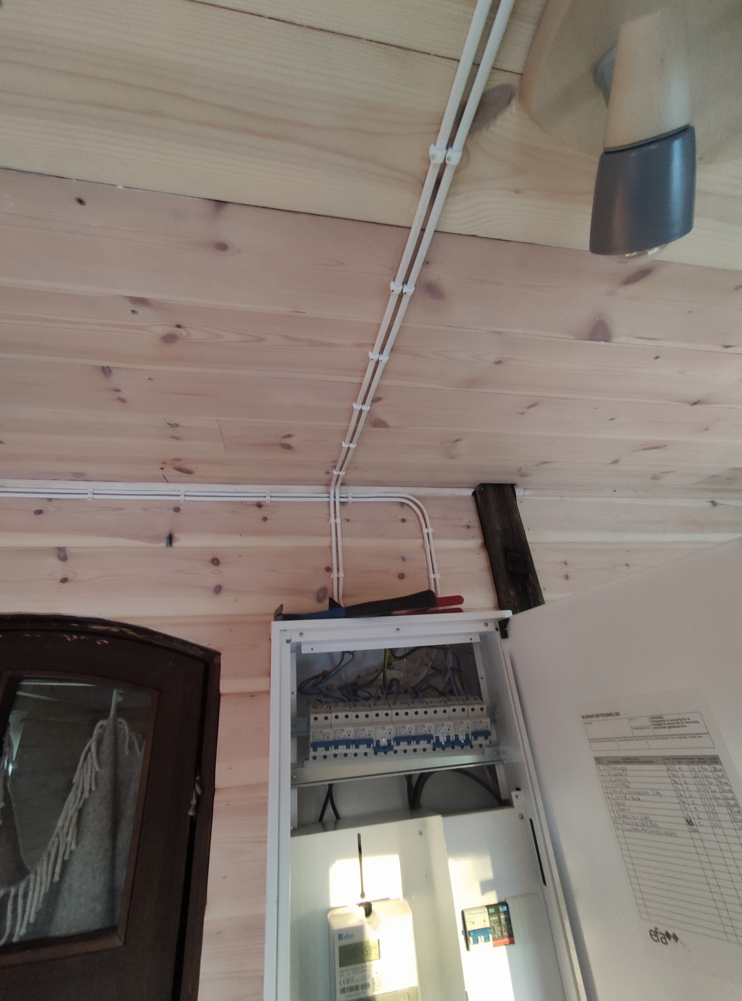 Rozdzielnica elektryczna z podłączonymi przewodami w drewnianym domu, widok z dołu. Widoczne zabezpieczenia i schemat instalacji.