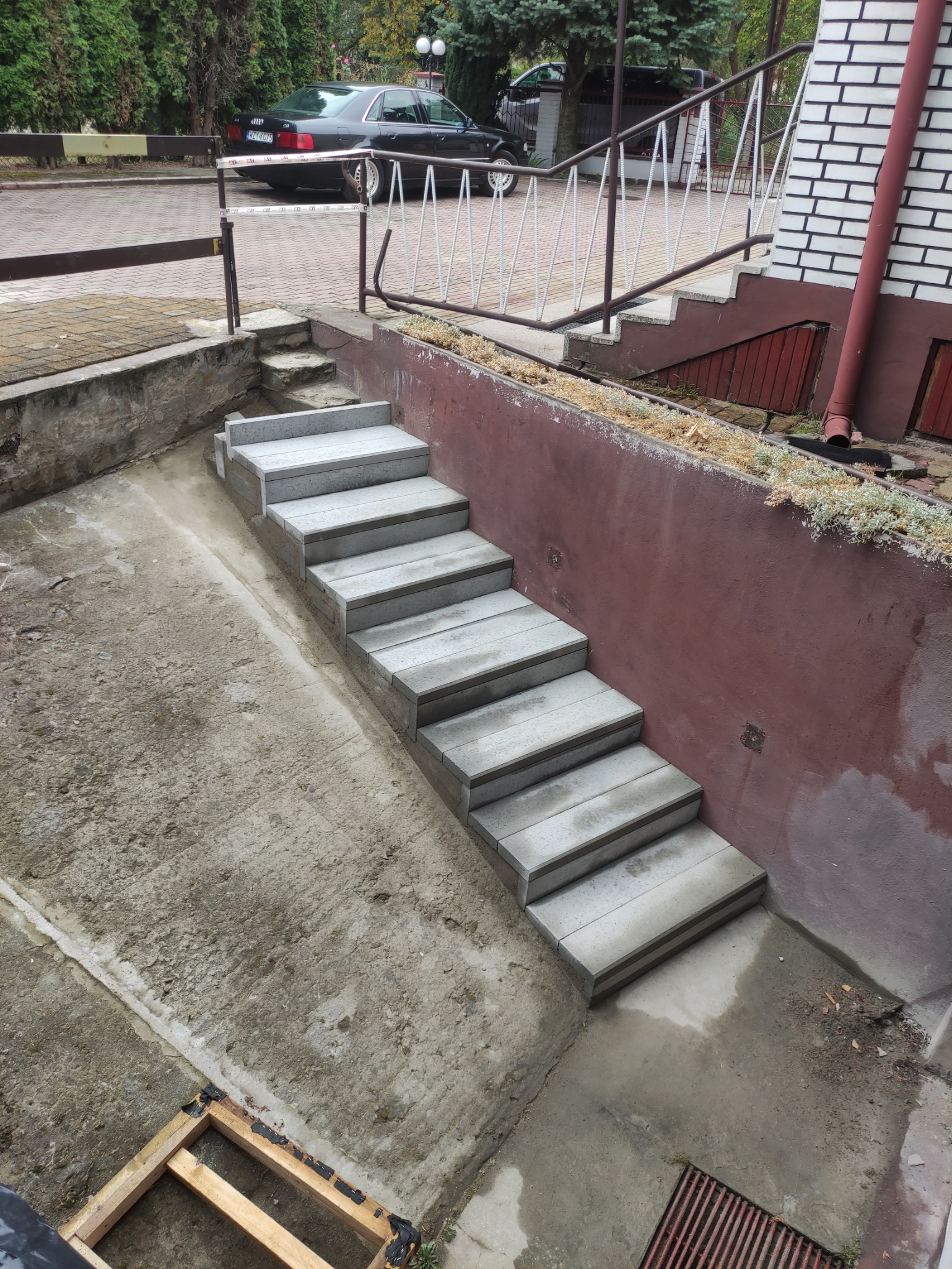Nowe, szare schody betonowe prowadzą w dół, kontrastując z czerwoną ścianą i elementami zieleni. W tle widoczny budynek i samochód.