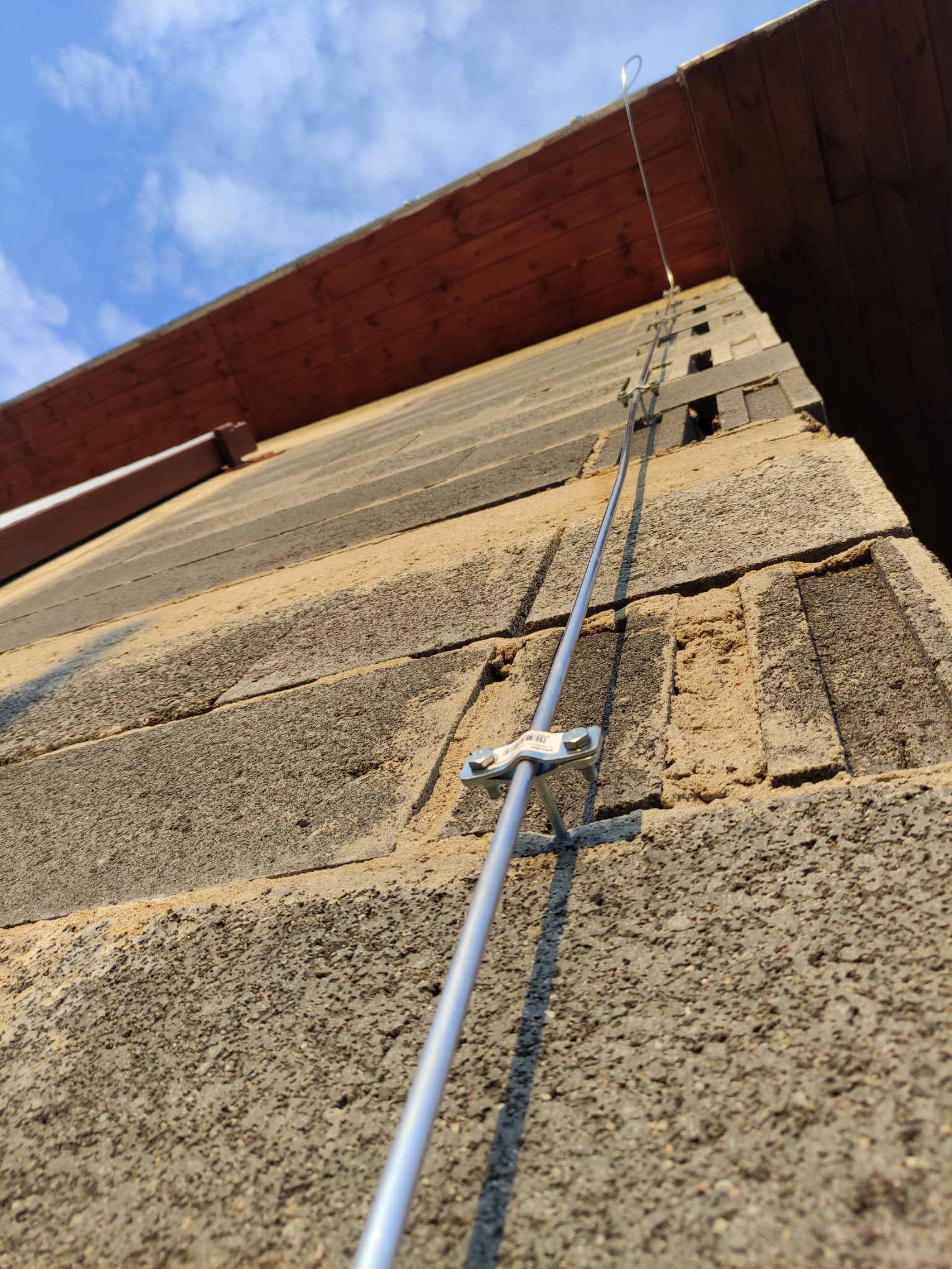 Zewnętrzna ściana budynku z zamontowanym systemem odgromowym, widoczne uchwyty mocujące i przewód. Ujęcie z dołu, niebo w tle.