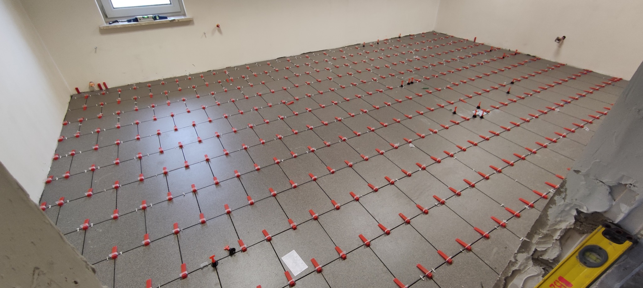Świeżo ułożone, szare płytki podłogowe z systemem poziomowania (czerwone klipsy) w jasnym pomieszczeniu. Widoczny poziomica przy ścianie.