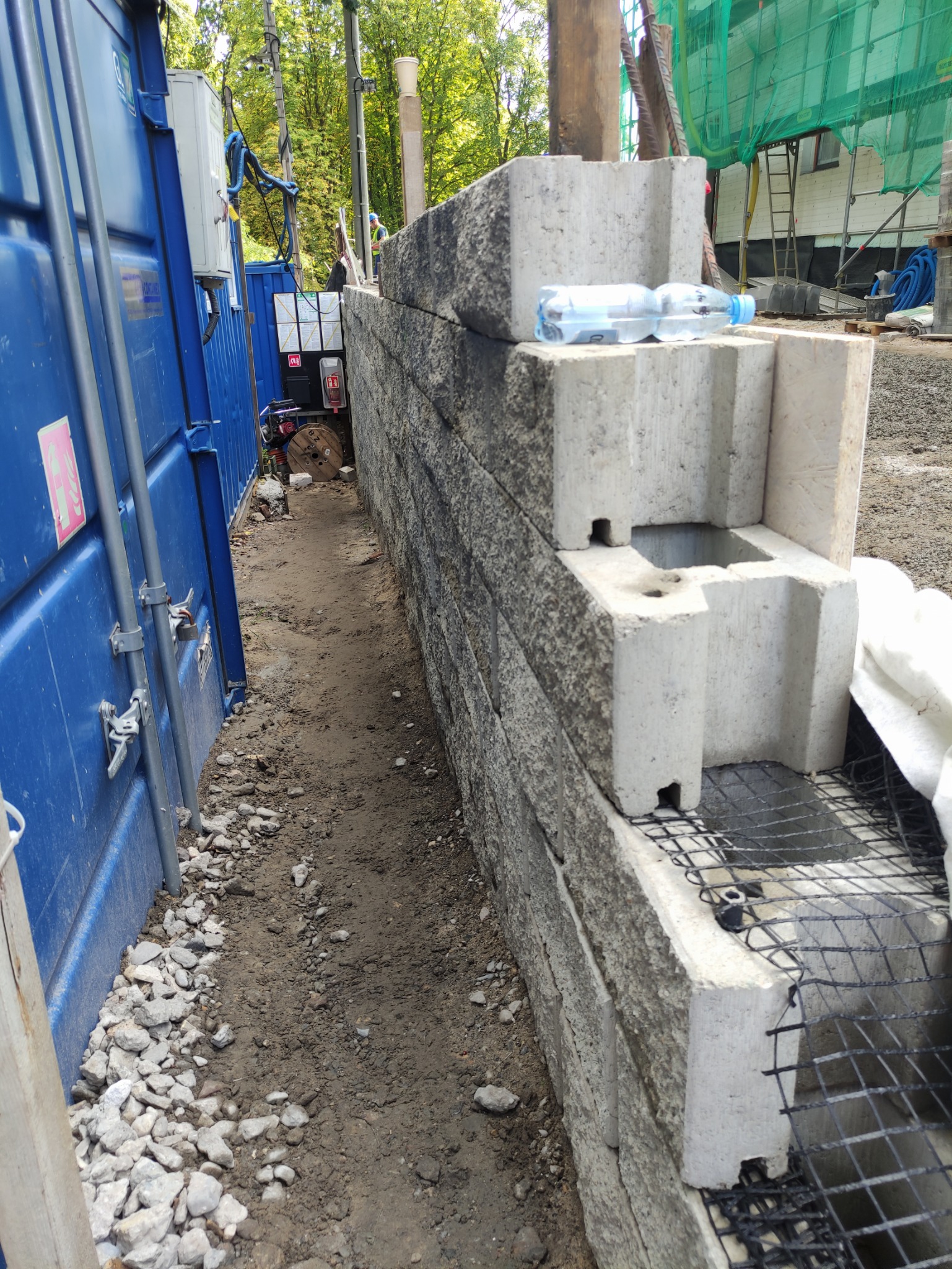 Wznoszenie muru oporowego z betonowych bloków. Widoczne elementy konstrukcyjne, siatka zbrojeniowa i butelka wody na szczycie muru. Prace budowlane w toku.