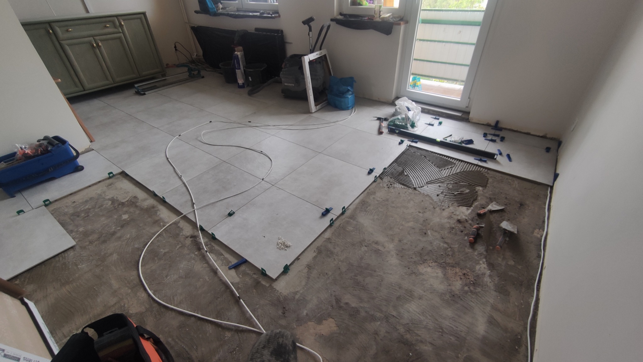 Układanie jasnoszarych płytek podłogowych w pokoju. Częściowo położone płytki, widoczny klej i narzędzia glazurnicze. W tle meble i okno balkonowe.