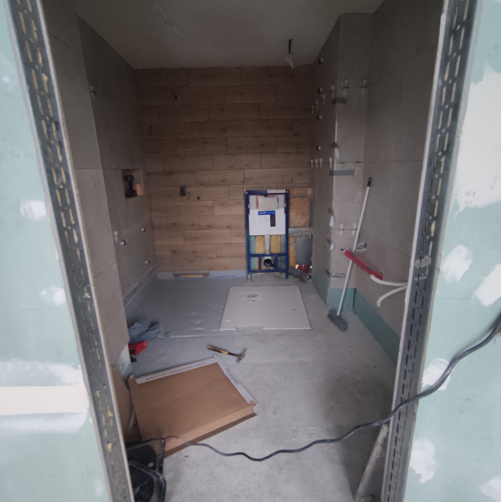 Łazienka w trakcie remontu: widoczne płytki imitujące drewno, stelaż podtynkowy WC, narzędzia i materiały budowlane. Prace wykończeniowe w toku.