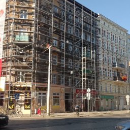 LDV - Budynek w rusztowaniach podczas renowacji elewacji, widoczny fragment odnowionej części, ulica z przechodniami i pojazdami w słoneczny dzień.