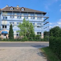 Wielopiętrowy budynek w trakcie renowacji, otoczony rusztowaniami i zielenią, widoczny z poziomu ulicy w słoneczny dzień. Częściowo pomalowana fasada.