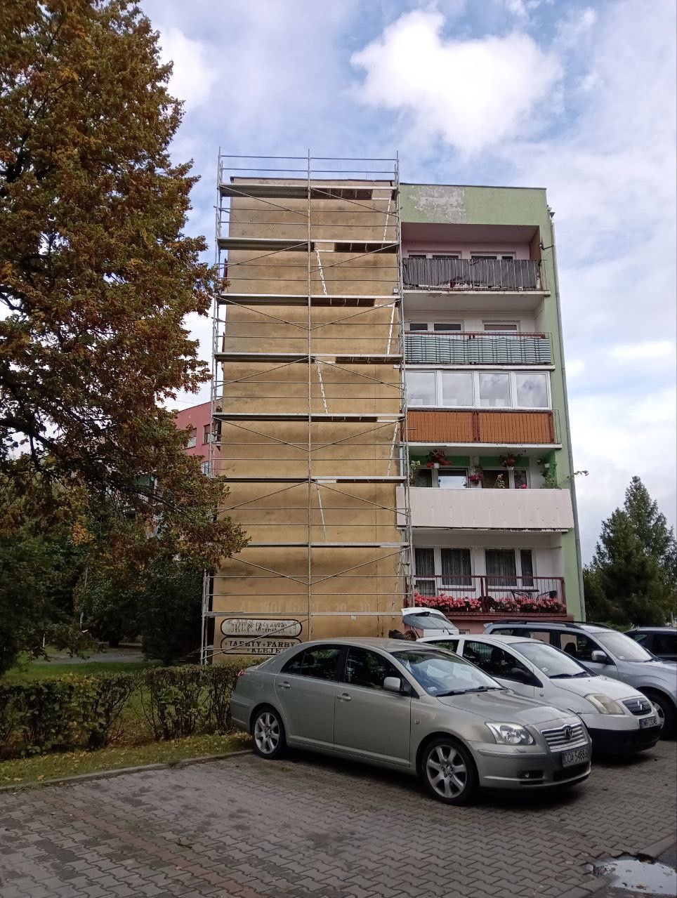 Ocieplanie bloku mieszkalnego ze styropianem, widoczne rusztowania, samochody na parkingu i drzewo z jesiennymi liśćmi. Prace na wysokości.