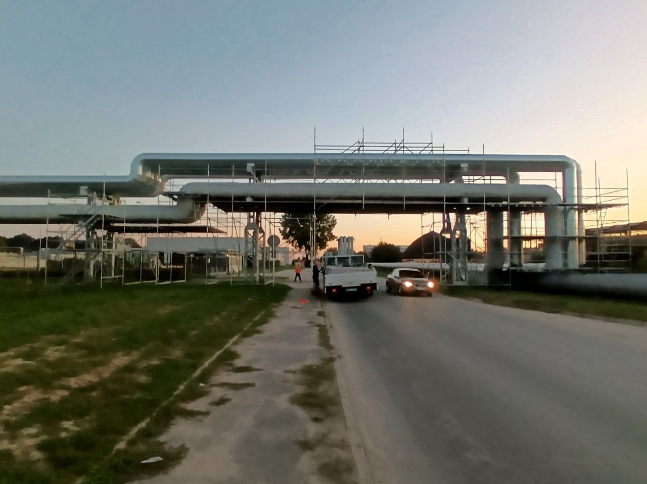 Rozległa instalacja rurociągowa na podporach, otoczona rusztowaniami, z pojazdami w pobliżu, sfotografowana o zmierzchu.