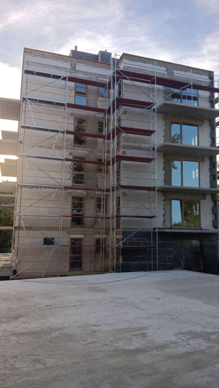 Budynek w trakcie budowy z rusztowaniami na elewacji. Widoczne okna i fragmenty konstrukcji balkonów. Prace na wysokości.