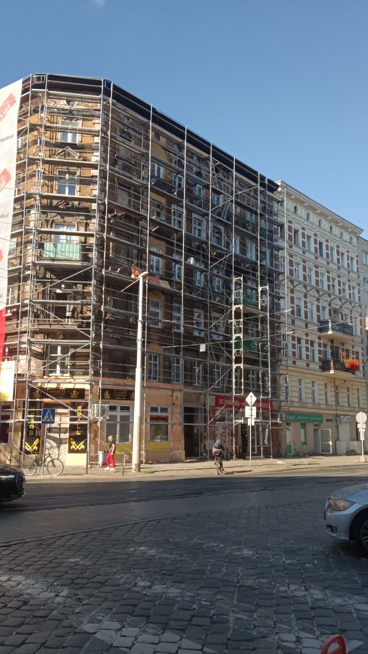 Budynek w rusztowaniach podczas renowacji elewacji, widoczny fragment odnowionej części, ulica z przechodniami i pojazdami w słoneczny dzień.