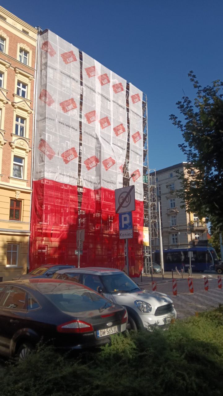 Budynek w rusztowaniach, osłonięty siatką z logo firmy KABE, częściowo w barwach flagi Polski. Prace wysokościowe w mieście.