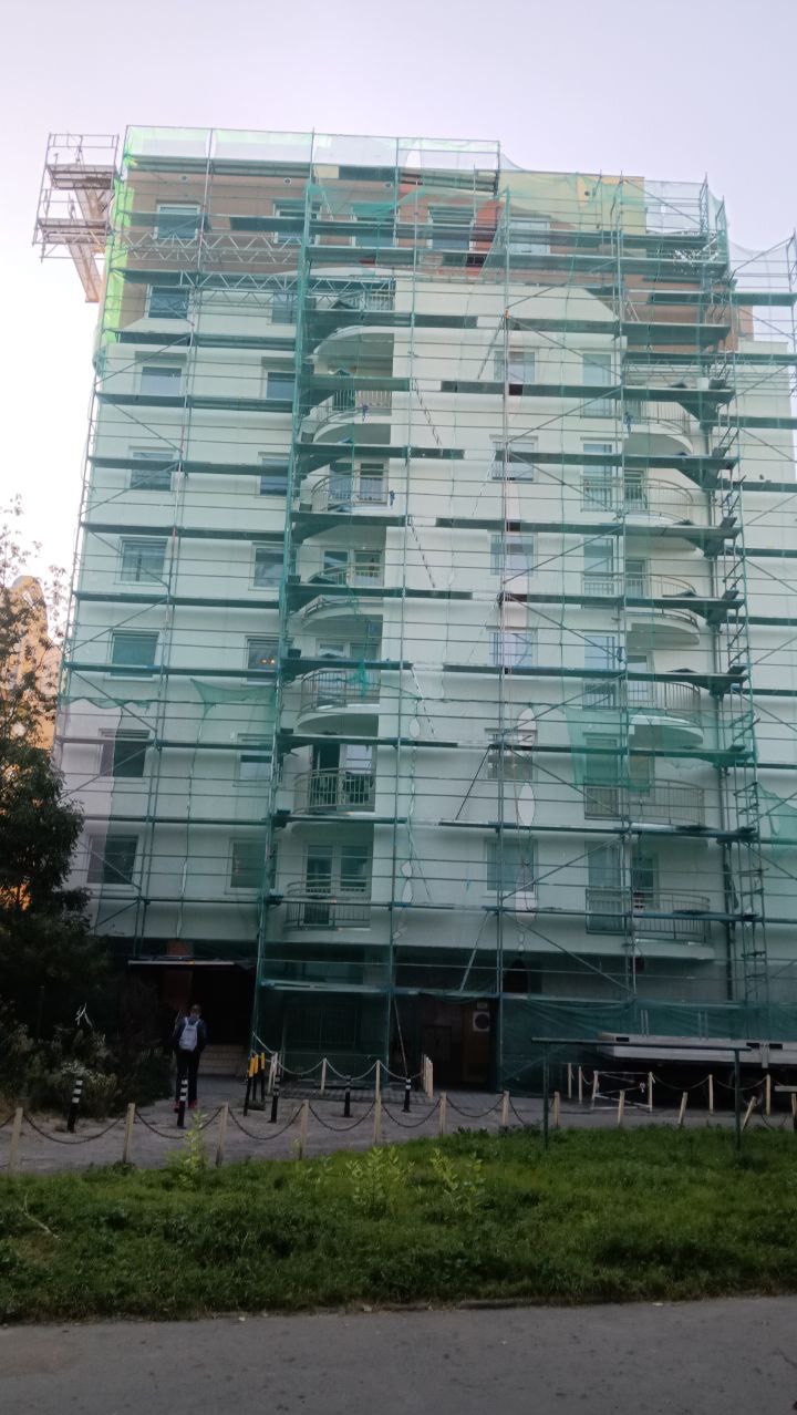 Wysoki budynek mieszkalny w trakcie remontu, osłonięty rusztowaniami i siatką zabezpieczającą we Wrocławiu. Widoczny fragment chodnika i zieleni.