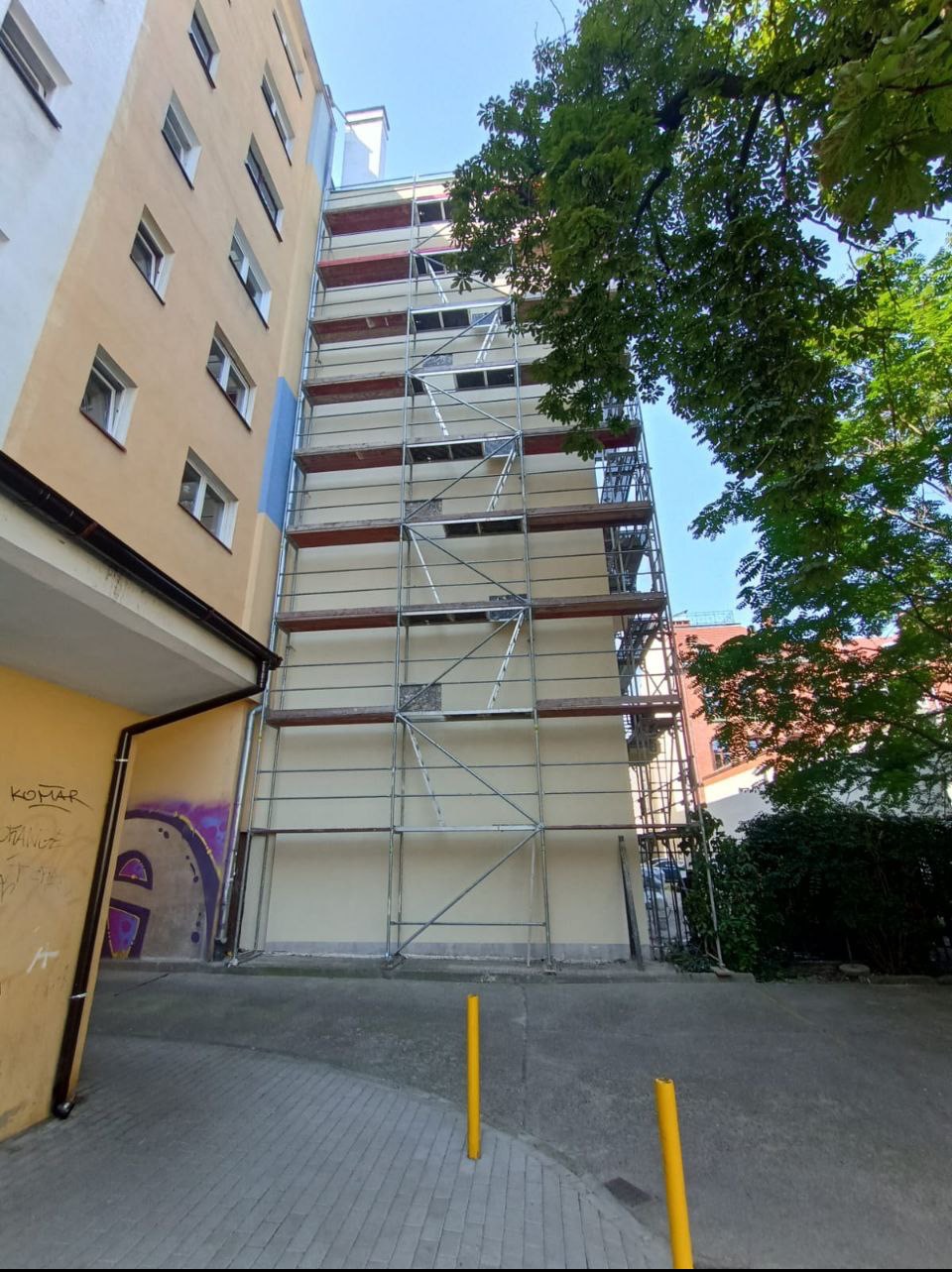 Wysoki budynek w trakcie prac remontowych, osłonięty rusztowaniami. Widoczne fragmenty elewacji w trakcie renowacji, zieleń drzew w tle i żółte słupki zabezpieczające.