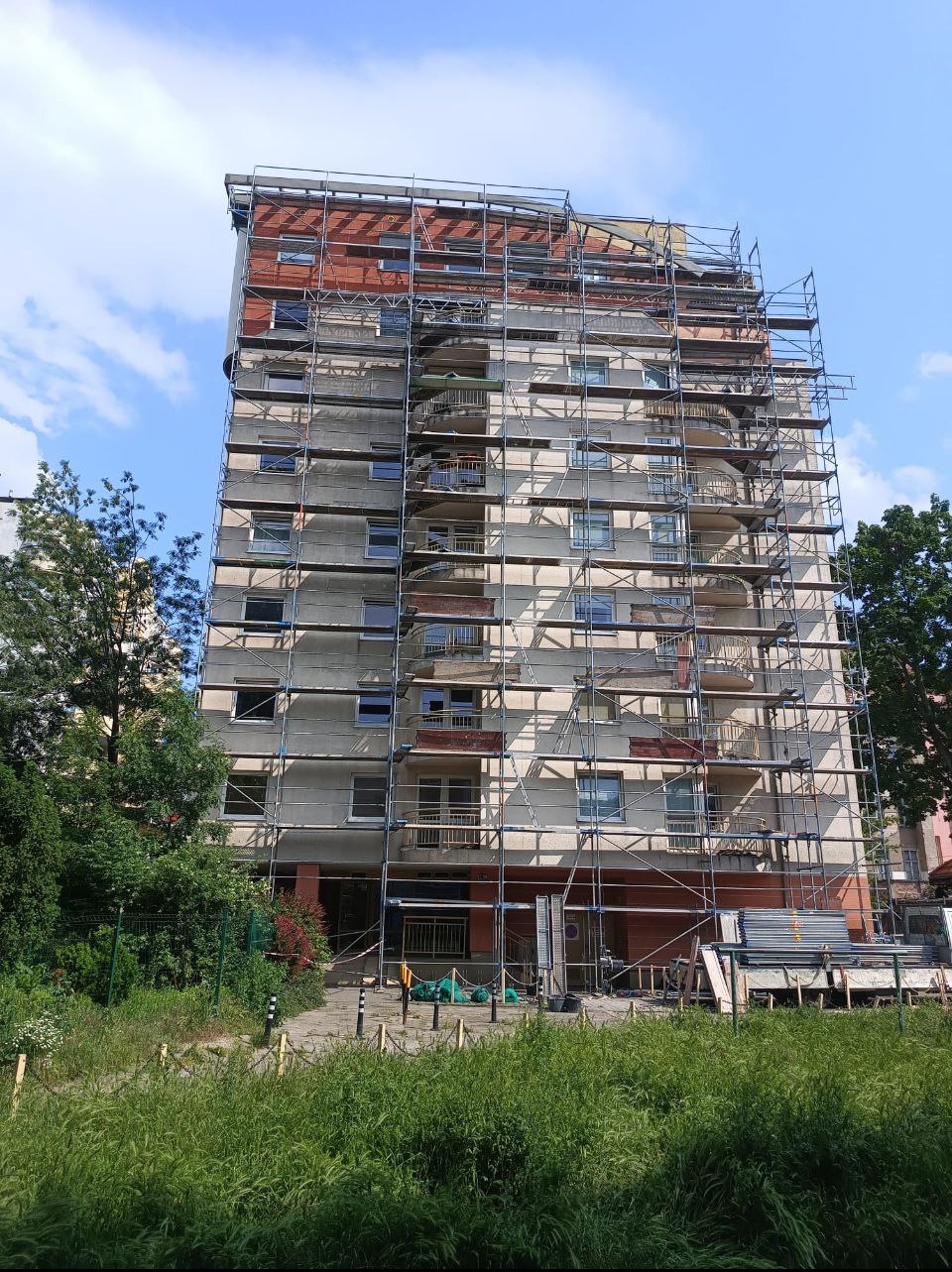 Wysoki budynek mieszkalny w rusztowaniach, przygotowywany do prac na wysokościach, z widocznymi elementami elewacji i zielenią w otoczeniu.