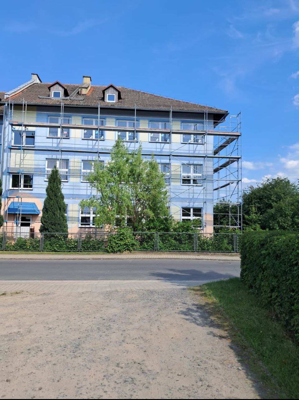 Wielopiętrowy budynek w trakcie renowacji, otoczony rusztowaniami i zielenią, widoczny z poziomu ulicy w słoneczny dzień. Częściowo pomalowana fasada.