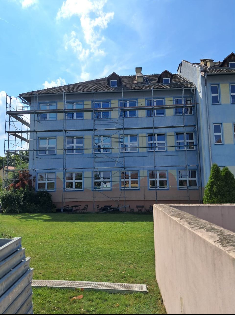 Budynek w trakcie prac elewacyjnych, z rusztowaniem na tle błękitnego nieba. Widoczny fragment trawnika i element ogrodzenia.