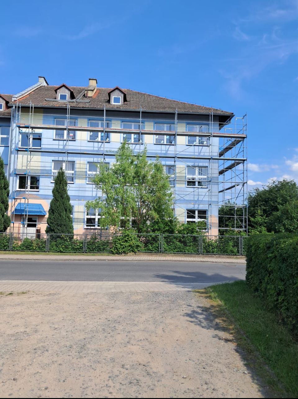 Budynek wielorodzinny w trakcie prac elewacyjnych. Widoczne rusztowania, błękitna fasada i elementy dekoracyjne. Prace na wysokościach w słoneczny dzień.